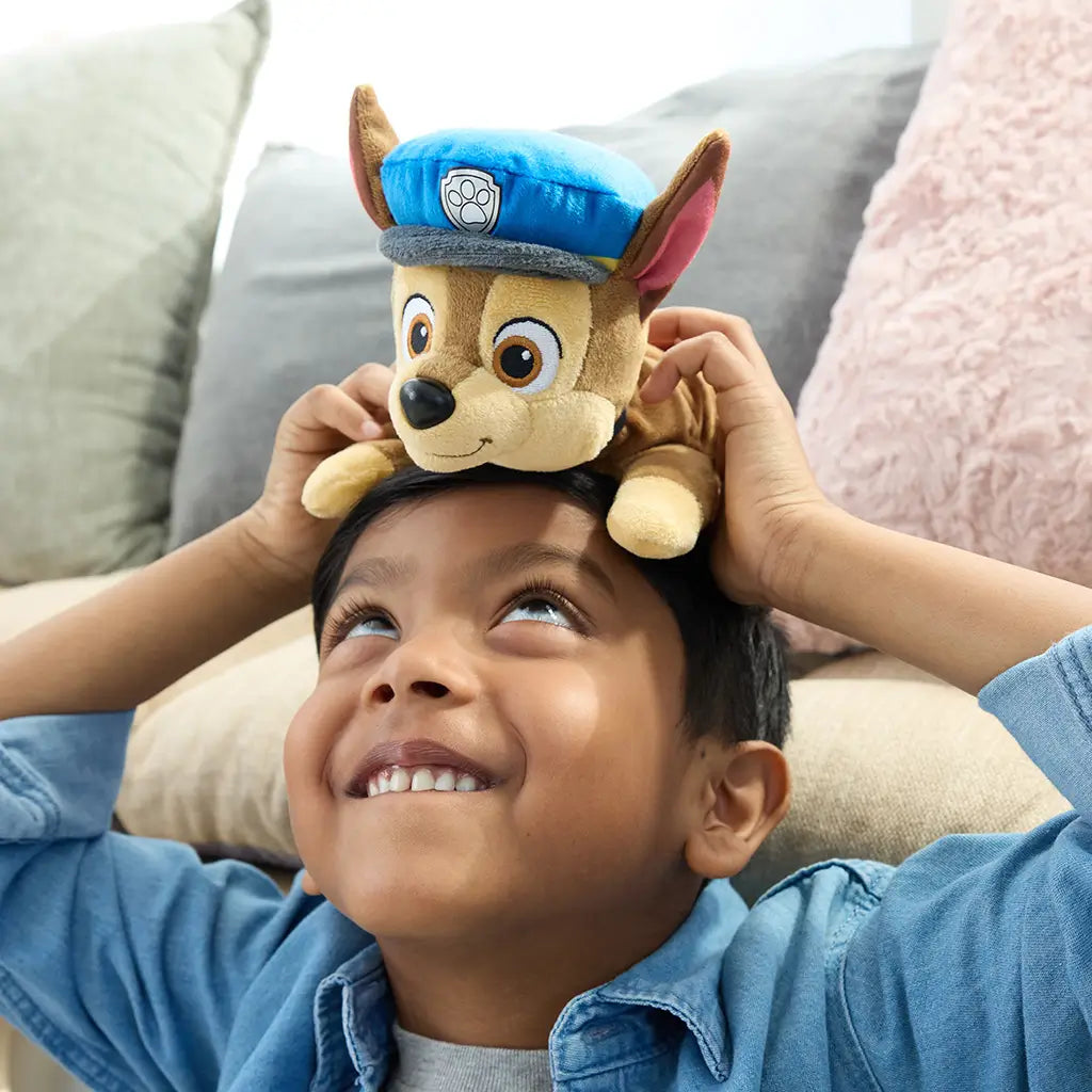 Paw Patrol Chase plush toy in brown and tan soft fabric with blue police hat, small rounded dog shape placed playfully on child’s head in bright indoor living room setting
