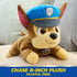 Paw Patrol Chase plush toy in brown and tan soft fabric with blue police hat, small rounded dog shape posed lying forward on couch in a bright indoor living room setting