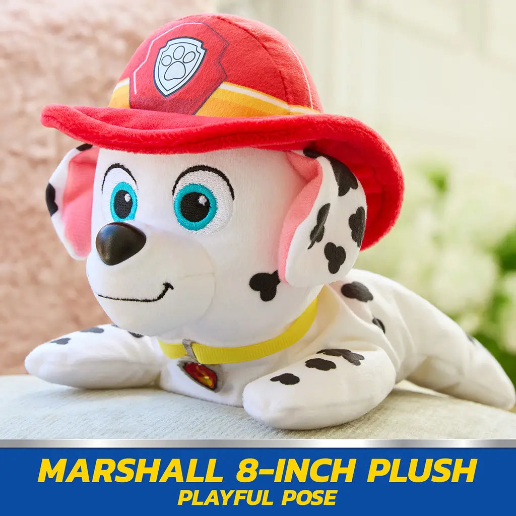 Paw Patrol Marshall plush toy in white and black spotted soft fabric with red firefighter hat, small rounded dog shape posed lying forward on couch in bright indoor living room