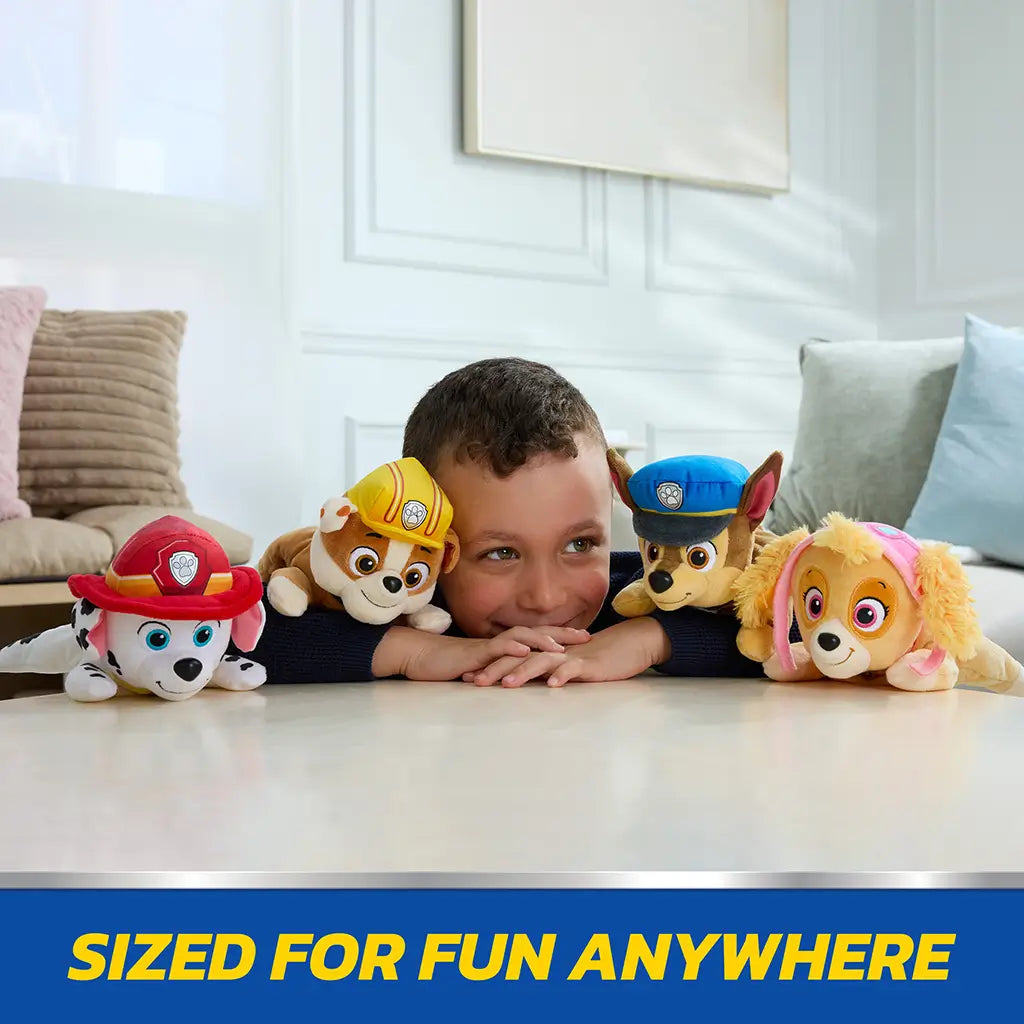 Paw Patrol plush toys in bright multicolor soft fabric including Rubble, Chase, Skye, and Marshall, small rounded dog figures arranged around smiling child on floor in a modern living room