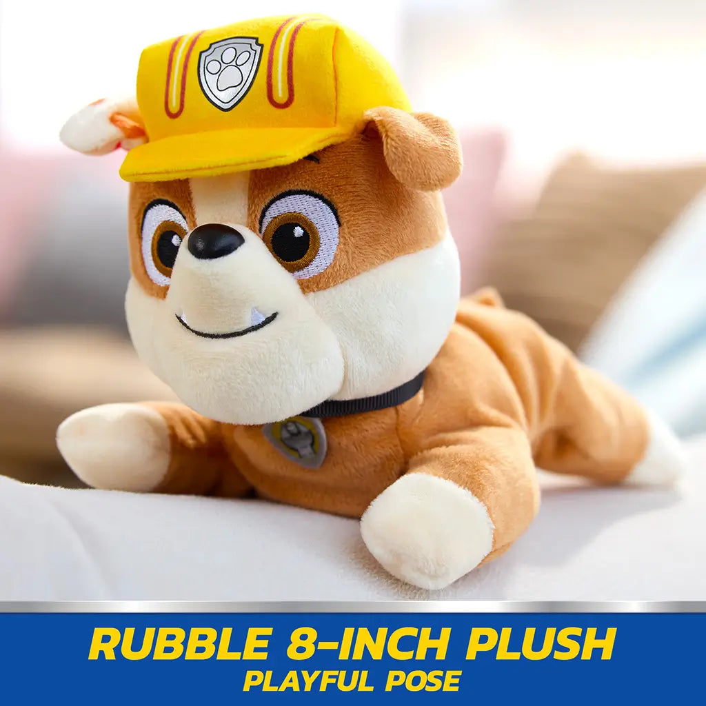 Paw Patrol Rubble plush toy in tan and cream soft fabric with yellow helmet, small rounded bulldog shape posed lying forward on couch in bright indoor living room setting