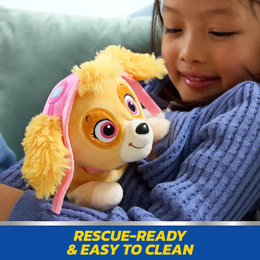 Paw Patrol Skye plush toy in tan and cream soft fabric with pink aviator hat and floppy ears, small rounded dog shape designed for cuddling, held by smiling child on couch indoors