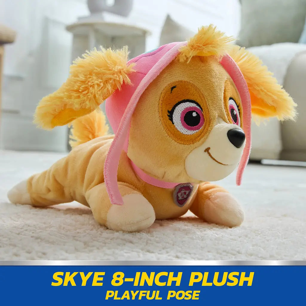 Paw Patrol Skye plush toy in tan and pink soft fabric with floppy ears and aviator hat, small rounded dog shape posed lying forward on carpet in bright indoor living room setting
