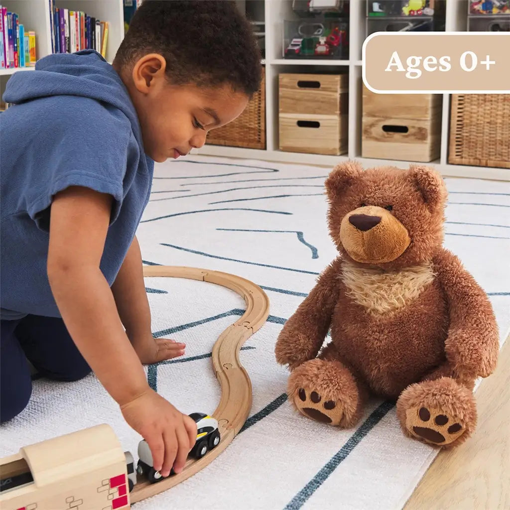 GUND Slumbers teddy bear plush in medium brown fuzzy fabric with plump seated bear form, embroidered nose and paw pads, shown beside a child playing with a toy train on a rug to emphasize ages zero plus playtime.