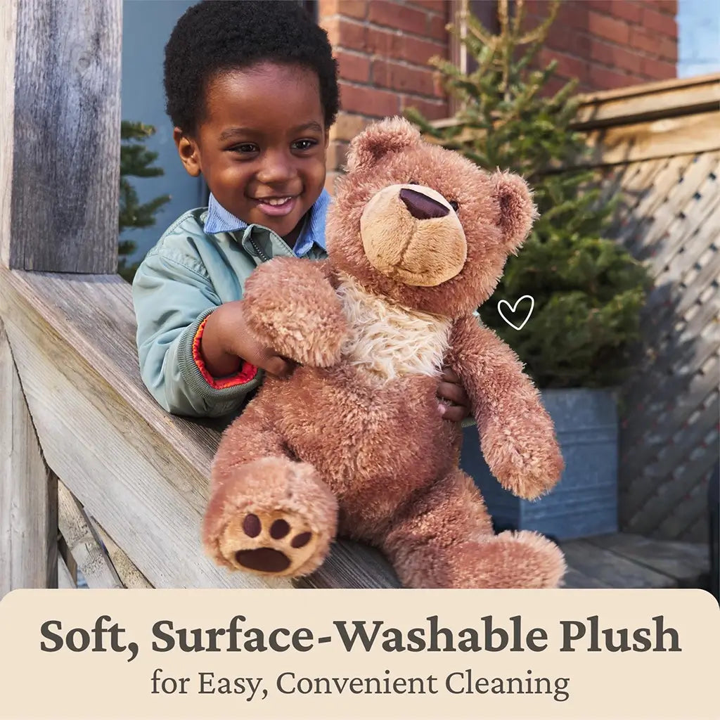 GUND Slumbers teddy bear plush in warm brown soft fabric with fluffy texture, rounded seated bear shape, embroidered nose and paw pads, shown outdoors with a child to highlight surface-washable, easy-care design.