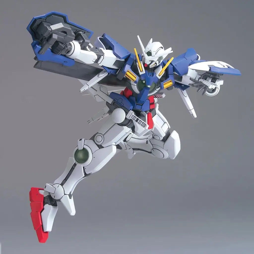 Gundam Exia GN-001 HG 1/144 model in blue, white, and red plastic with articulated limbs, posed mid-air with arms extended and shield raised, displayed on a plain gray studio backdrop.