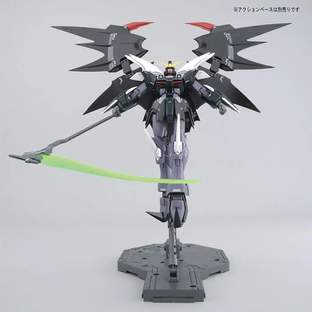 Bandai Gundam Deathscythe Hell front view in black and gray plastic with red chest vents and yellow V-fin, large batlike wings extended, gripping a green beam scythe while mounted on a stand against white background.