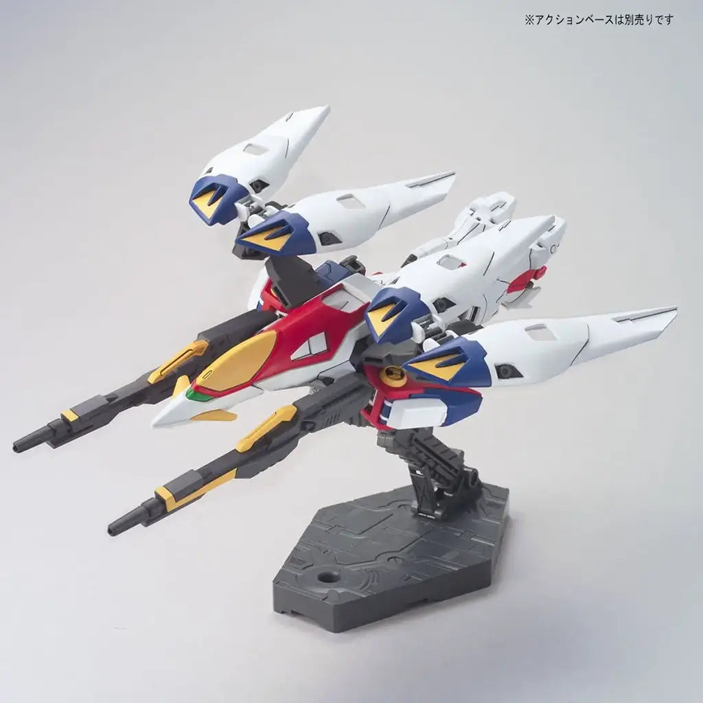 Bandai Wing Gundam Zero model kit in white, blue, red, and yellow plastic with jet mode form, featuring extended wings and mounted weapons, transforming mecha posed on a display stand against a neutral background