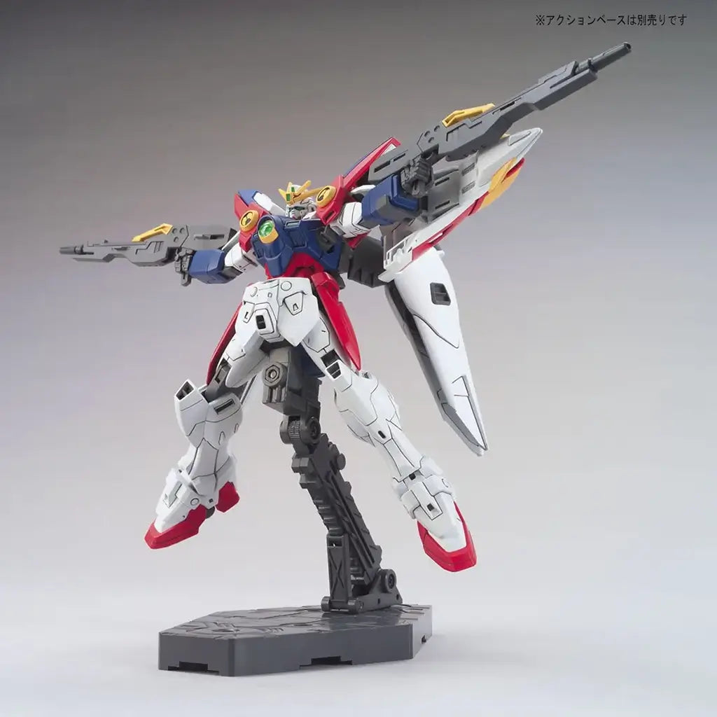 Bandai Wing Gundam Zero model kit in white, blue, red, and yellow plastic with humanoid robot form, featuring dual cannons and wing units extended, articulated mecha posed dynamically on a display stand background
