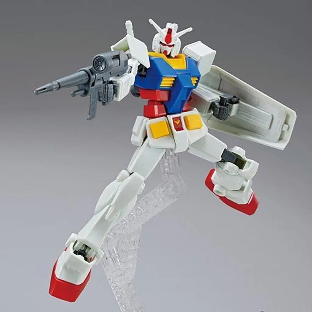 Gundam RX-78-2 model kit in white, blue, red, and yellow plastic with angular armor, posed mid-air holding a gray rifle and shield, displayed on a clear stand against a neutral gray studio background.