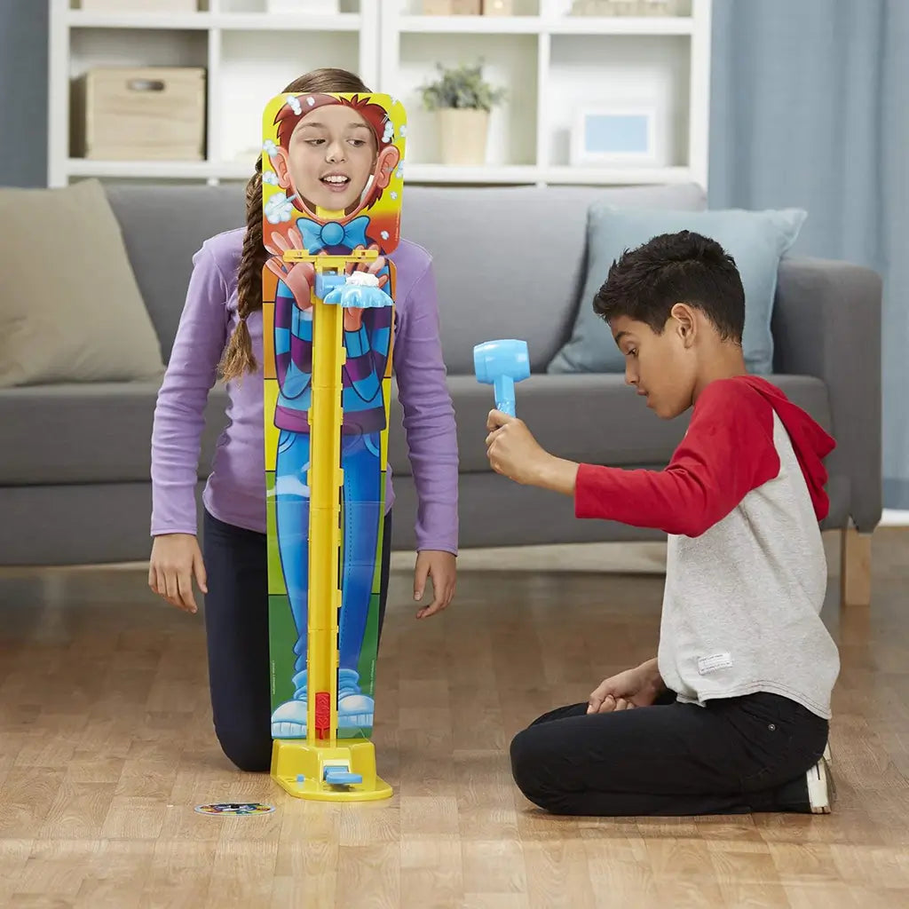 Two kids playing Pie Face Sky High game indoors, with one hitting the base using a mallet and the other standing behind the tall pie target ready to be “pied.”