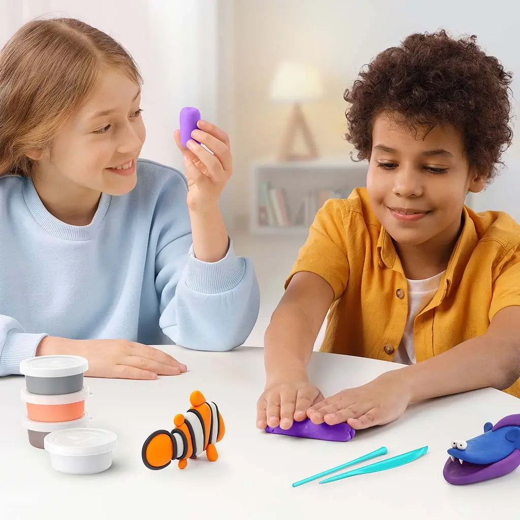 Two children crafting with the Hey Clay ocean set at a white table, shaping colorful clay pieces while completed sea creature figures and stacked tubs sit between them.
