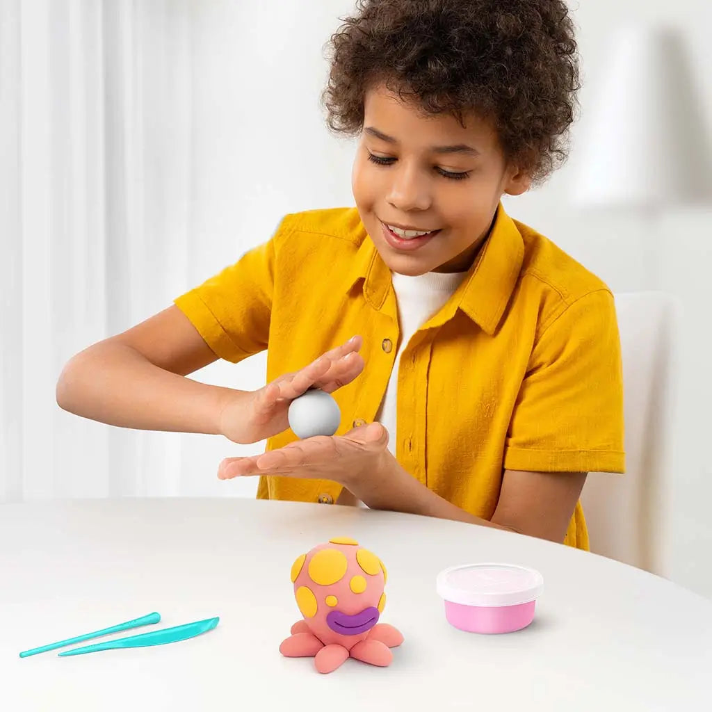 Child shaping a smooth clay ball from the Hey Clay ocean set at a white table, with a completed octopus figure, sculpting tools, and an open clay tub nearby.