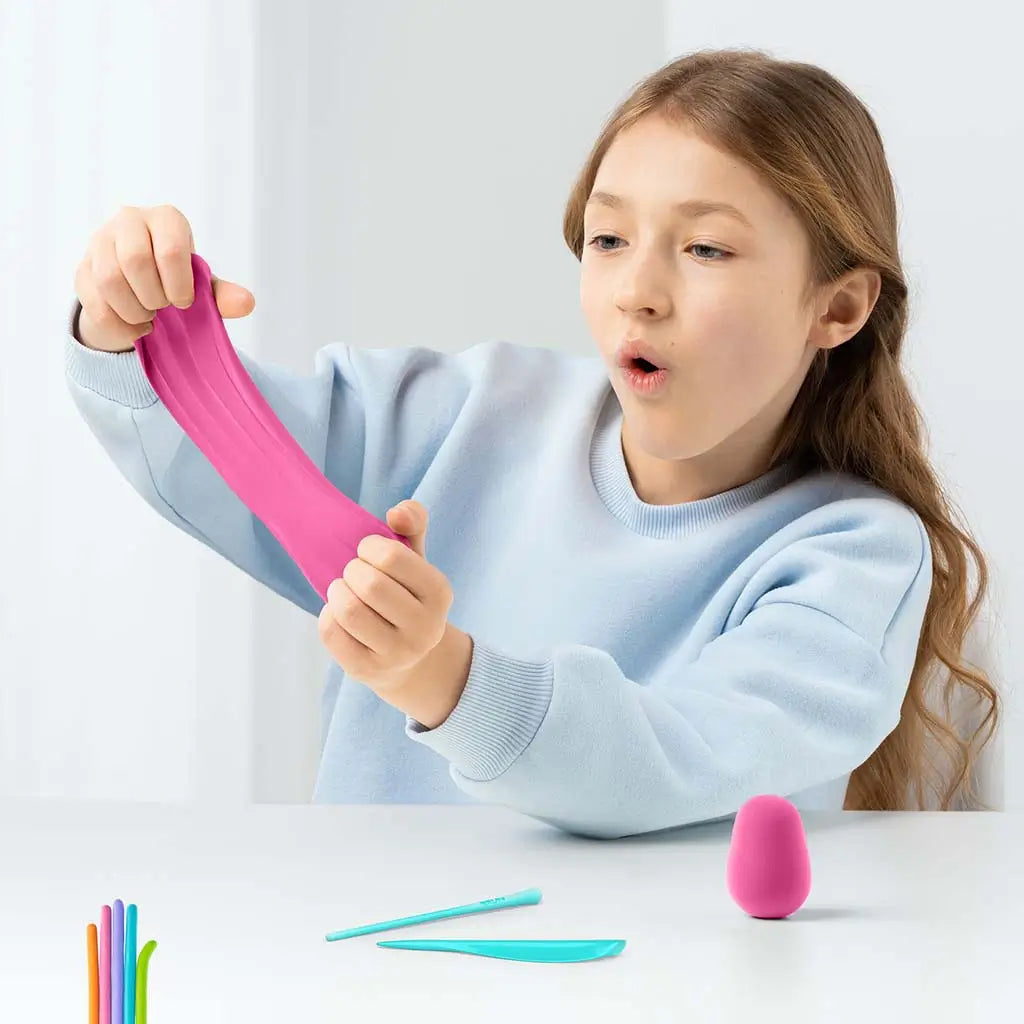 Child stretching bright pink Hey Clay material between both hands at a white table, with blue sculpting tools and a small shaped clay piece arranged nearby.