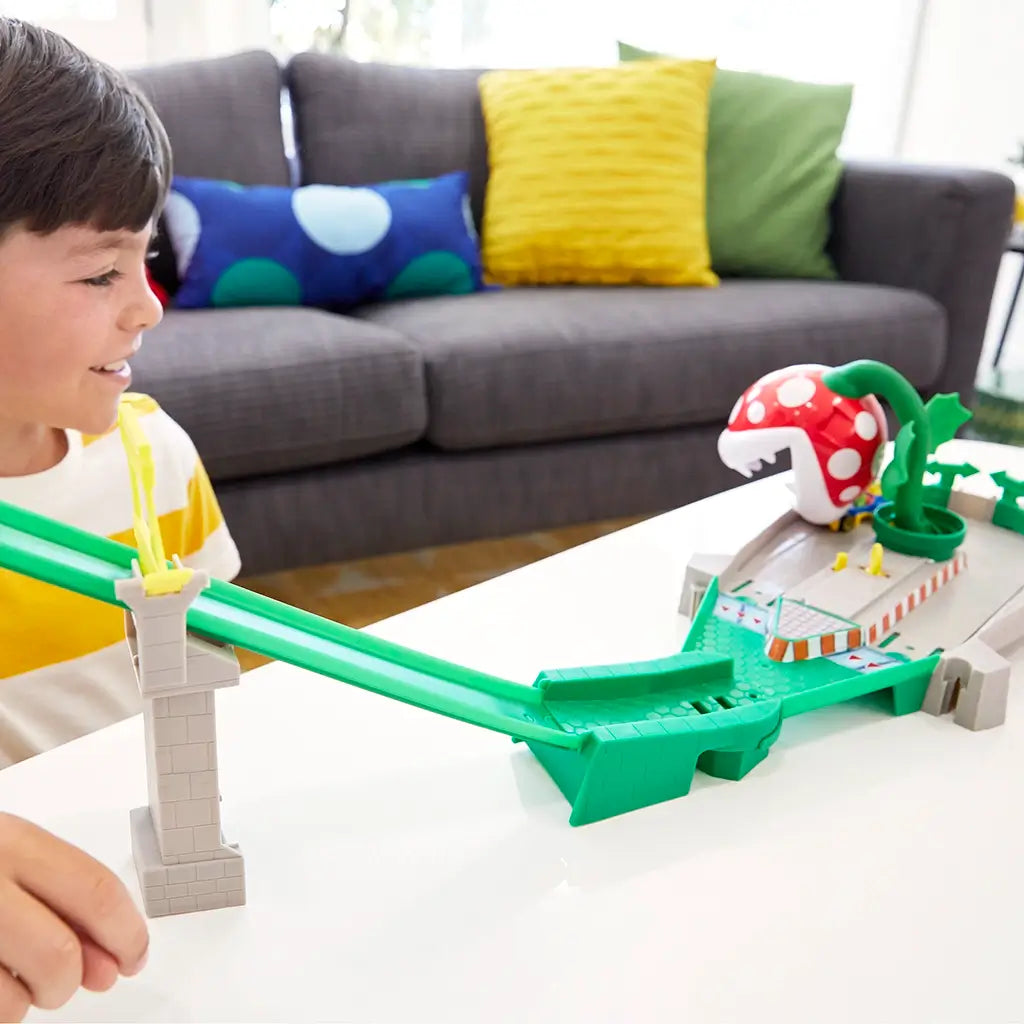 Hot Wheels Mario Kart track set with bright green plastic slide, yellow launcher, and large red Piranha Plant as a child smiles while playing in a modern living room.
