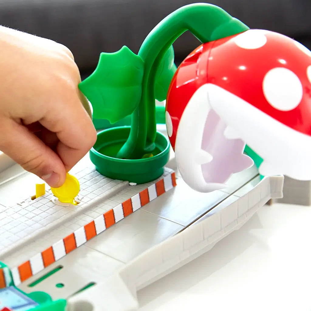Close-up of Hot Wheels Mario Kart track showing a hand placing a yellow coin near the large red Piranha Plant with white spots and green leaves on the playset’s base.