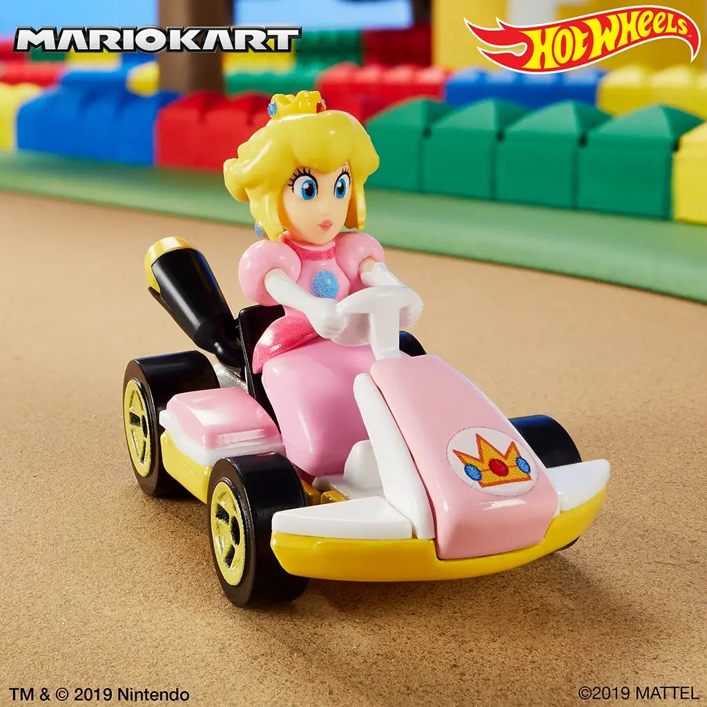 Hot Wheels Mario Kart Princess Peach kart in pink and yellow plastic racing across a toy track scene, showing the seated character figure, rolling gold wheels, and standard kart shape within a colorful playset setting.