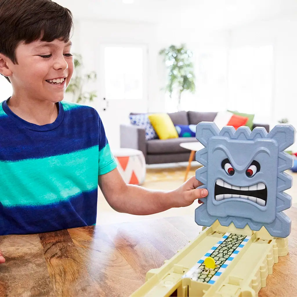 Smiling boy plays at a wooden table with the Hot Wheels Mario Kart Thwomp Ruins set, pressing the large gray Thwomp block above a tan race track decorated with castle-like textures.
