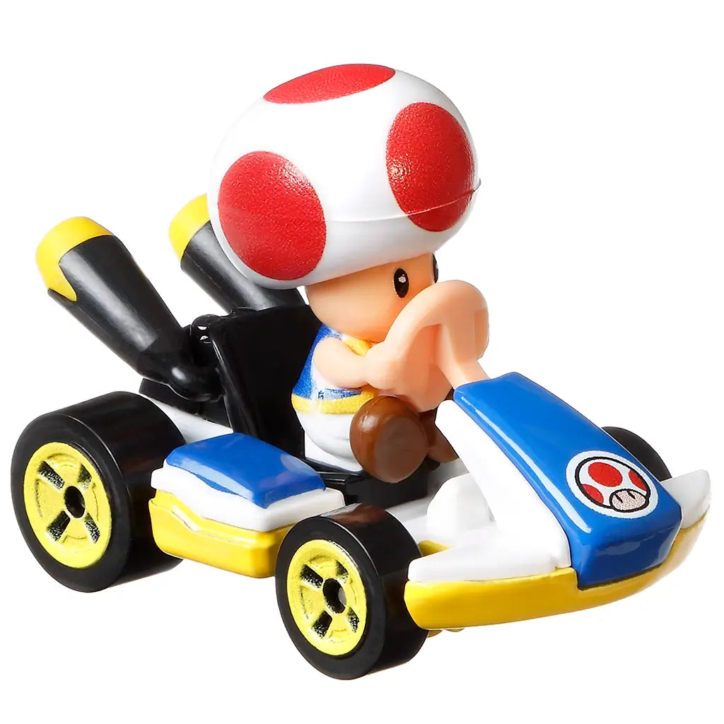 Hot Wheels Mario Kart Toad Standard Kart toy in white, blue, yellow, and black glossy plastic, featuring Toad seated at the wheel, mushroom emblem nose, gold wheels, and twin exhaust pipes on a clean white background.