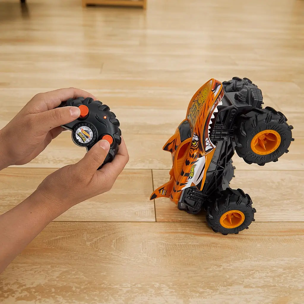 Orange and black Hot Wheels Tiger Shark RC monster truck with shark-like body and large treaded tires, shown performing a wheelie on a light wood floor while being remote-controlled.