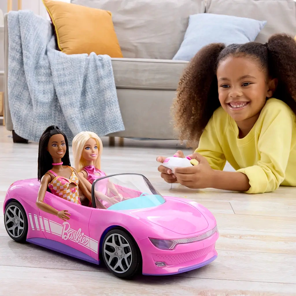 Smiling child plays indoors with Barbie RC convertible car in pink holding two Barbie dolls, using the included white remote control for interactive fun.