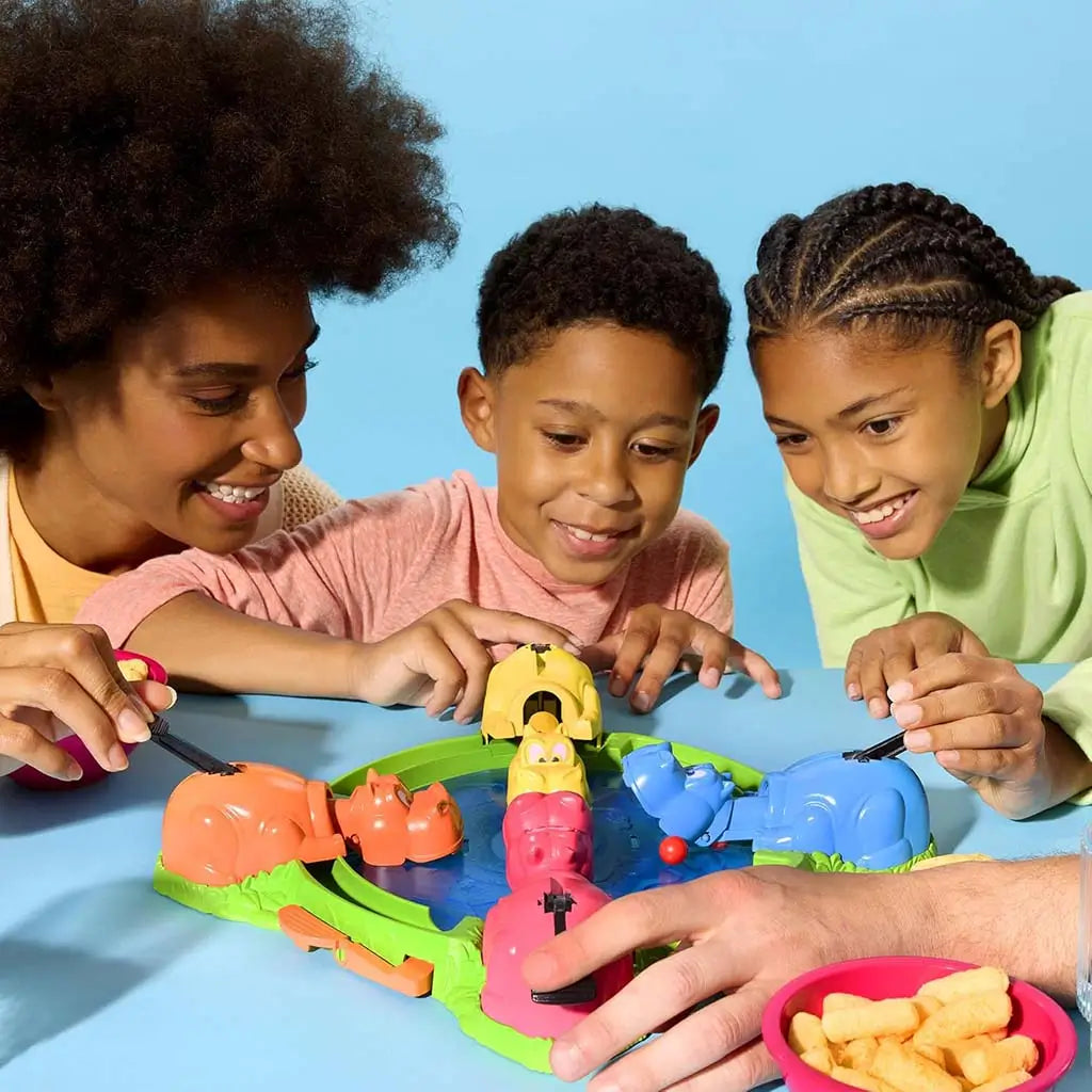 Hasbro Hungry Hungry Hippos family play moment with smiling kids leaning over the colorful green and blue board, pressing levers to help their hippos snap marbles in energetic competition.