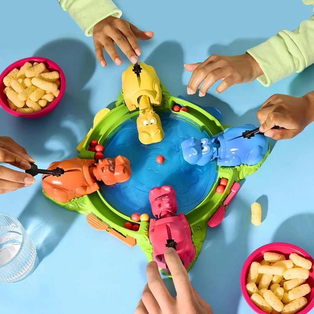 Hasbro Hungry Hungry Hippos on a blue tabletop with children’s hands reaching toward bright hippos as red marbles scatter across the board during lively snack-filled group play.
