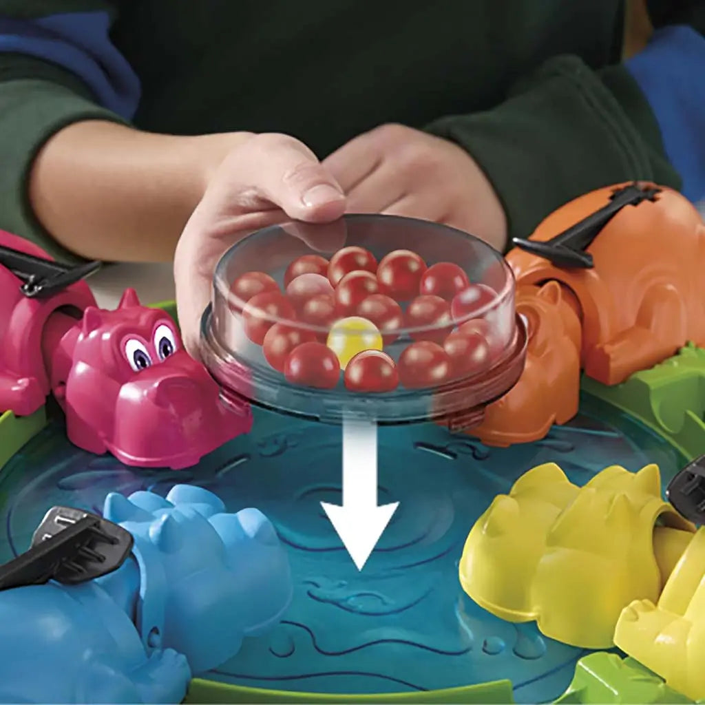 Hasbro Hungry Hungry Hippos top view with a bowl of red marbles being poured into the blue play pool while colorful hippos wait at the edges for fast-paced marble-chomping action.