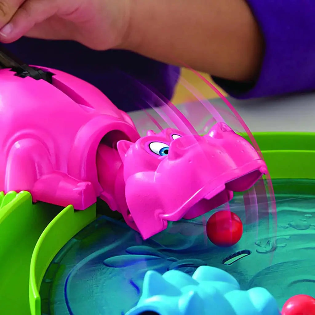 Hasbro Hungry Hungry Hippos reload moment with a pink hippo lunging toward a rolling red marble as a child presses the lever to trigger quick snapping motion on the glossy blue board.