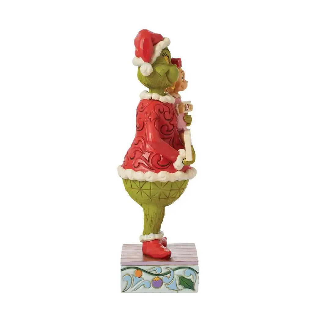Side view of Jim Shore's Grinch figurine holding Cindy Lou Who, wearing festive red attire and Santa hats, standing on a decorative base with scroll details.