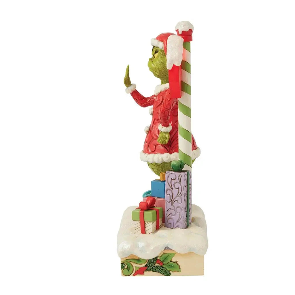 Alternate side view of Grinch LED figurine by Jim Shore, standing near colorful gift boxes and a striped holiday pole topped with a snowy stop sign.