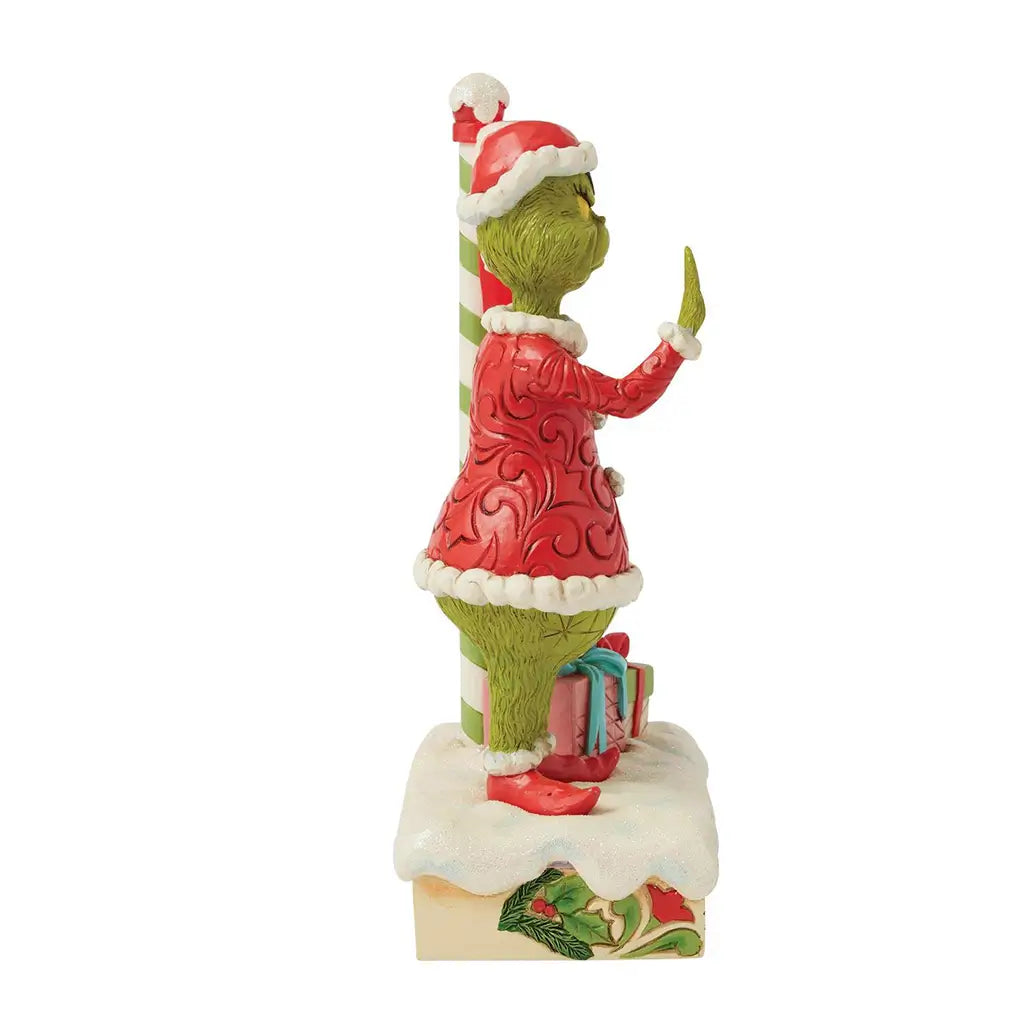 Side view of Jim Shore Grinch LED figurine in Santa outfit pointing near a striped pole with gifts and snow base, part of festive holiday décor.