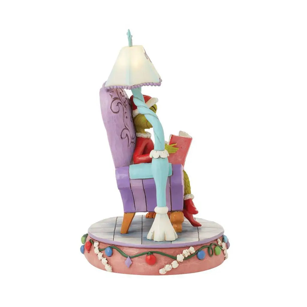 Side view of Jim Shore Grinch LED figurine seated in a purple chair reading a book under a glowing lamp, with Max the dog beside him in a Santa hat on a festive base.