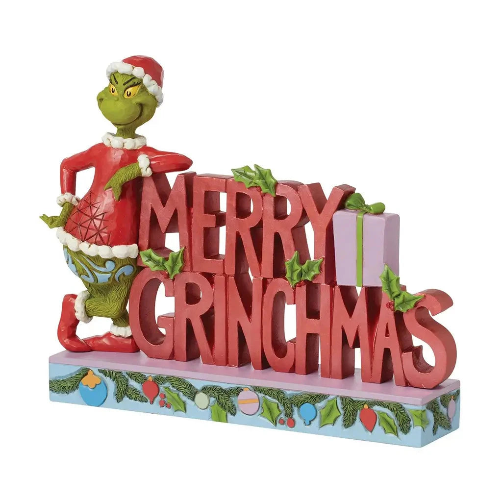 Angled view of Jim Shore "Merry Grinchmas" figurine featuring the Grinch posing next to stacked red holiday letters and festive holly accents.