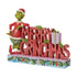 Angled view of Jim Shore "Merry Grinchmas" figurine featuring the Grinch posing next to stacked red holiday letters and festive holly accents.