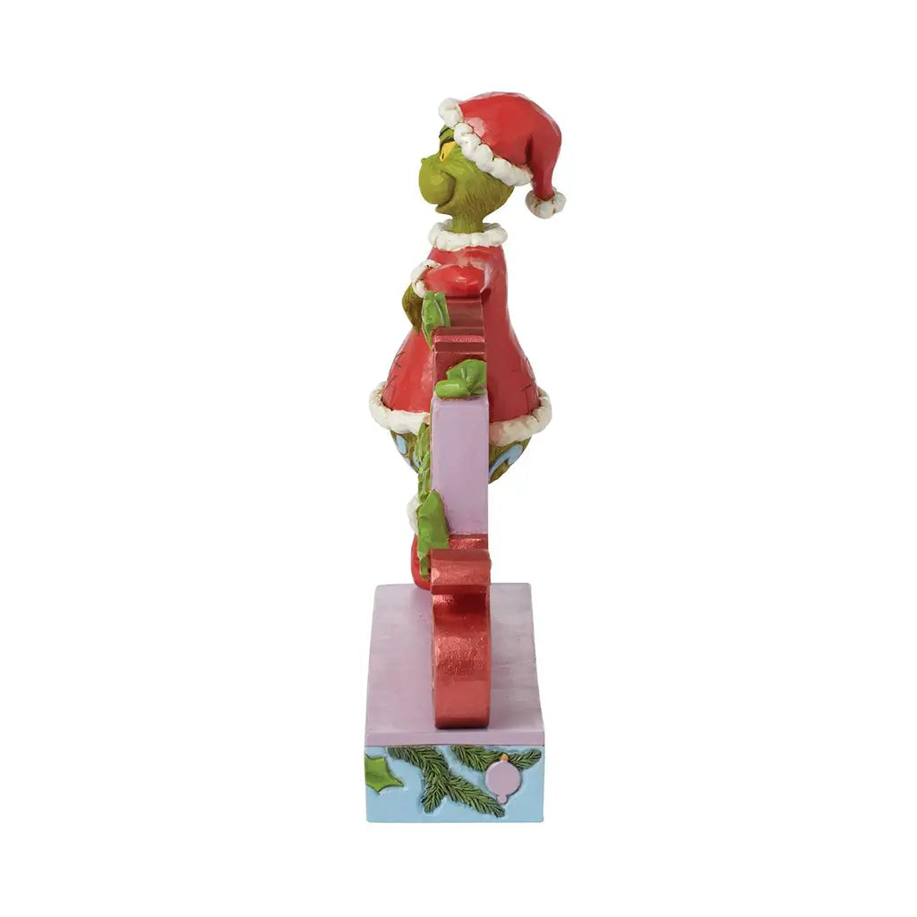 Side profile of Jim Shore "Merry Grinchmas" figurine showing the Grinch in Santa outfit leaning on the letters, with holiday detailing on the base.