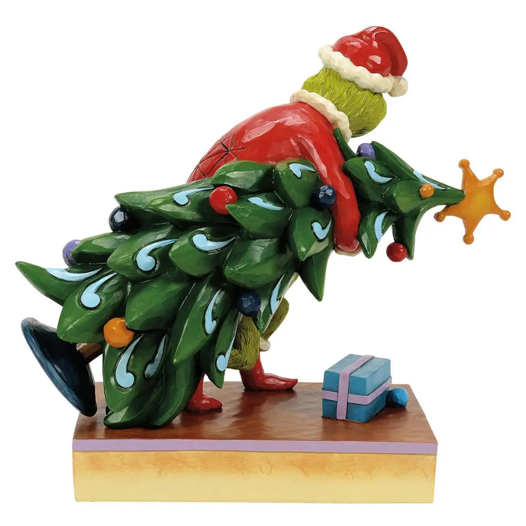 Back view of Jim Shore Grinch figurine in red Santa coat with white trim, holding decorated green Christmas tree on holly-trimmed base, against plain white background.