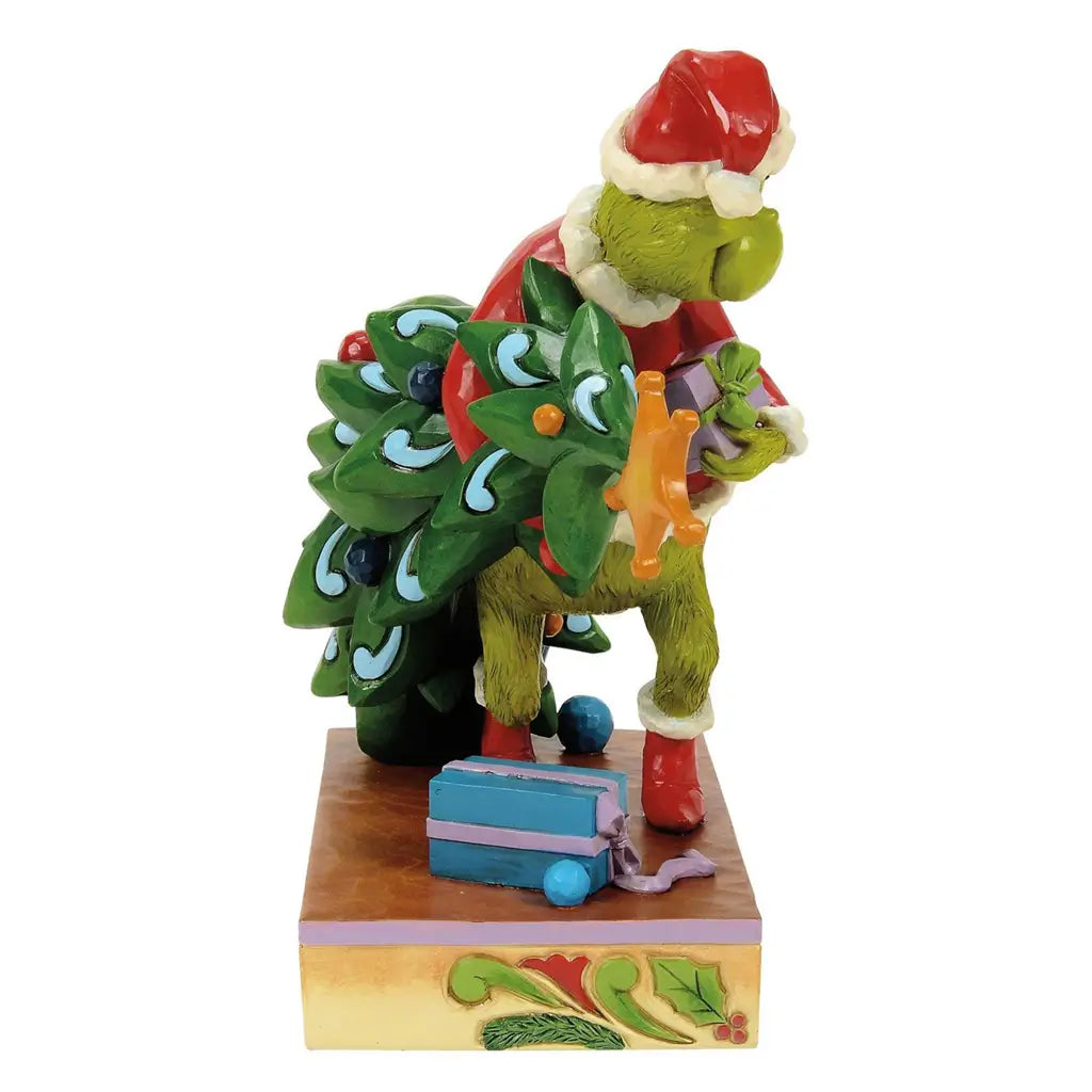 Side view of Jim Shore Grinch figurine in Santa outfit carrying Christmas tree with ornaments, stepping over presents on holly-trimmed base.