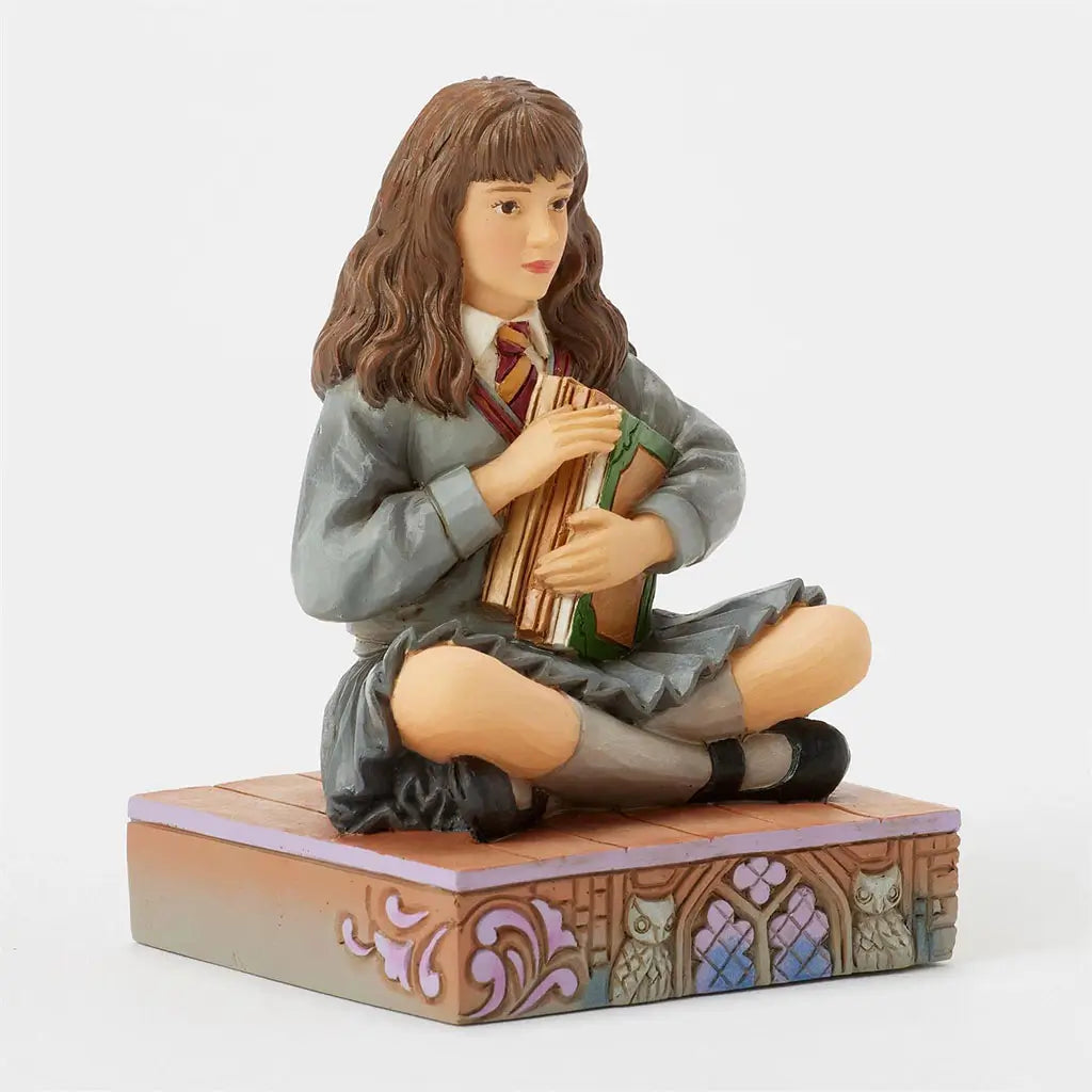 Jim Shore Harry Potter figurine of Hermione Granger, hand-painted in school uniform with Gryffindor tie, seated cross-legged holding a book atop a decorative stone base.