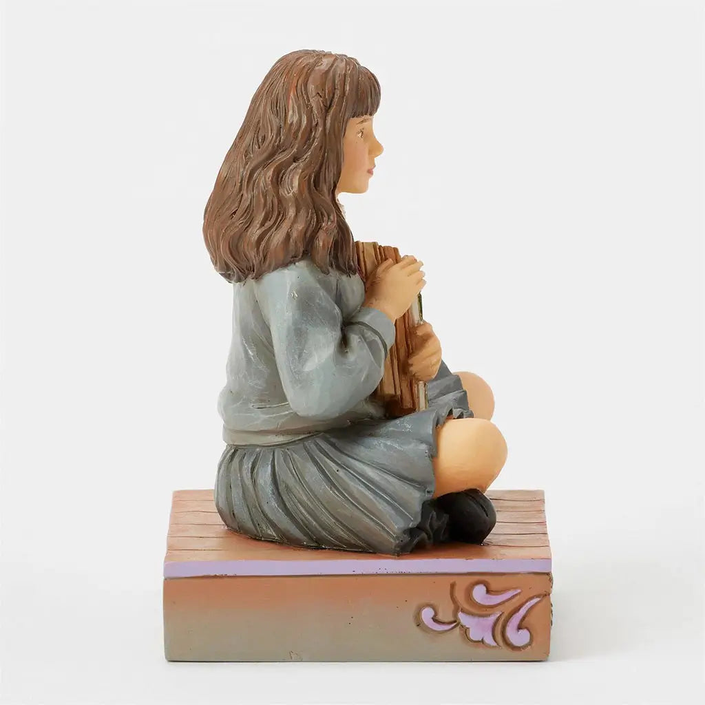 Left side view of Hermione Granger Jim Shore figurine, showing her brown wavy hair and grey pleated skirt while holding a detailed green and tan book in her lap.