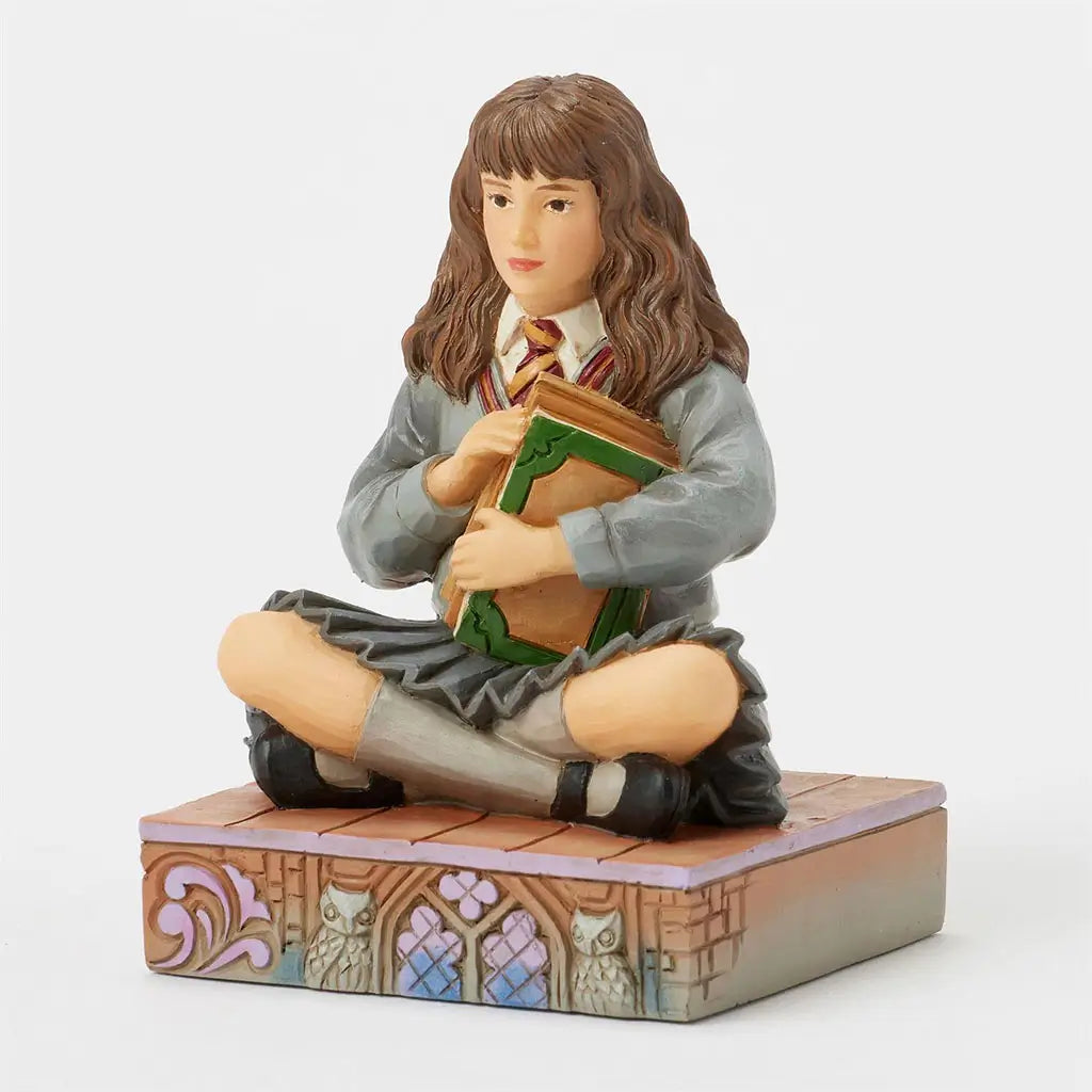 Right angled view of Hermione Granger Jim Shore figurine, showing textured carving style, green book cover details, and base featuring whimsical scroll and owl patterns.