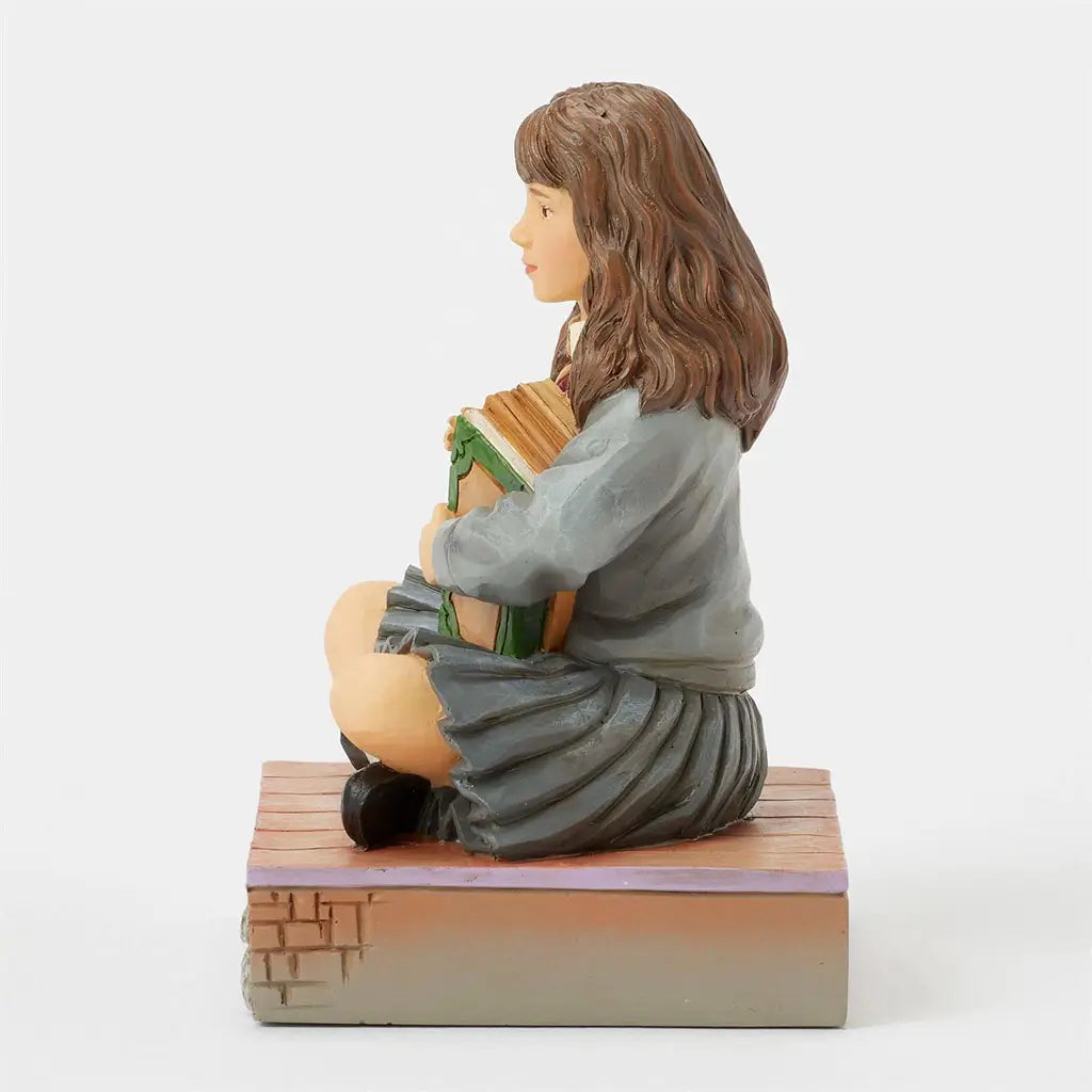 Right side view of Hermione Granger Jim Shore figurine, seated on a carved platform with painted brick details, book clutched to chest, skirt folds visible.