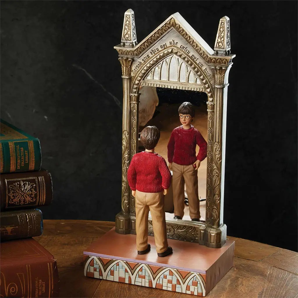 Lifestyle photo of Jim Shore Harry Potter Mirror of Erised figurine shows Harry gazing into golden gothic mirror, placed on wooden table beside vintage books.