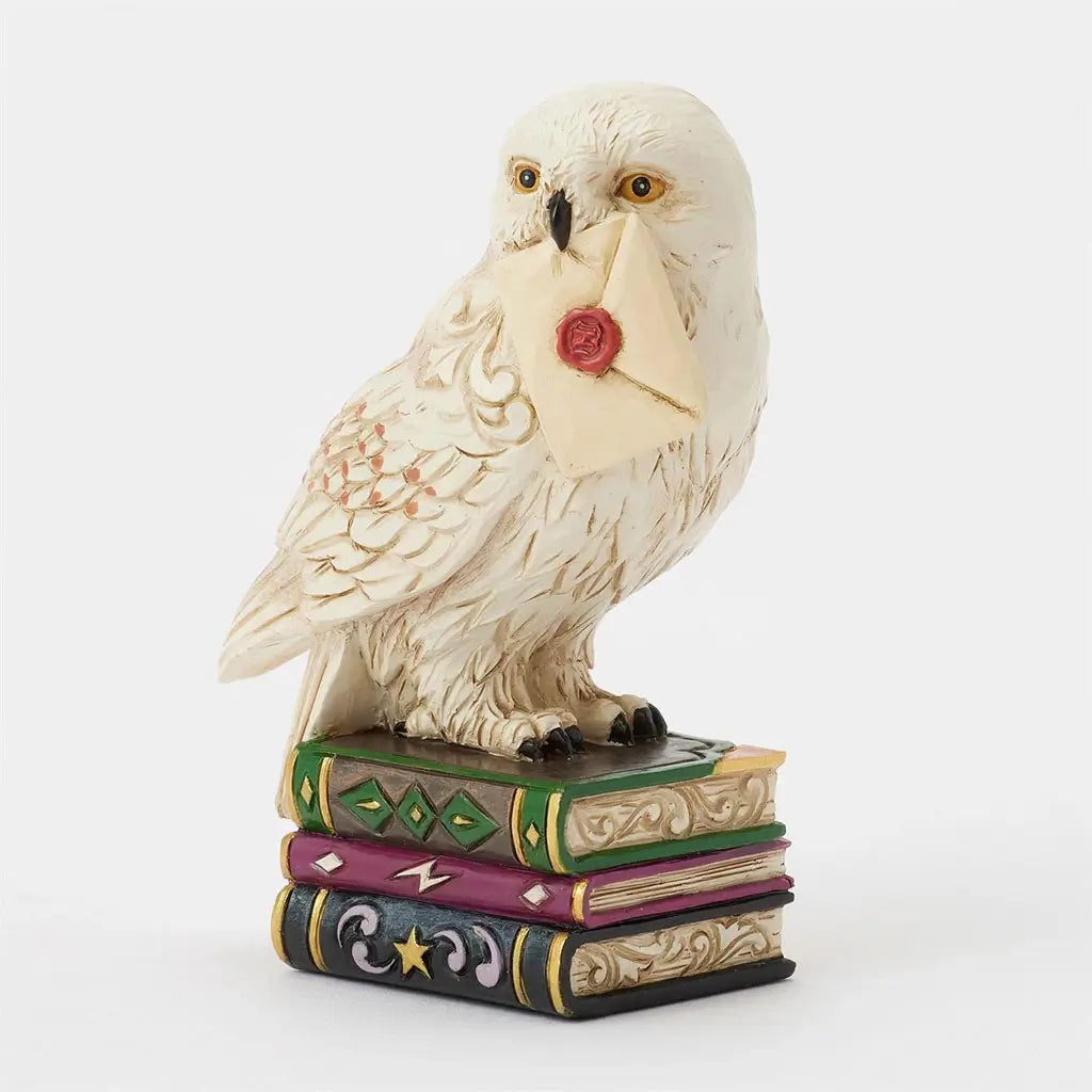 Angled Jim Shore Hedwig figurine shows snowy owl with golden eyes and feather detail, carrying Hogwarts letter while perched atop three colorful books.