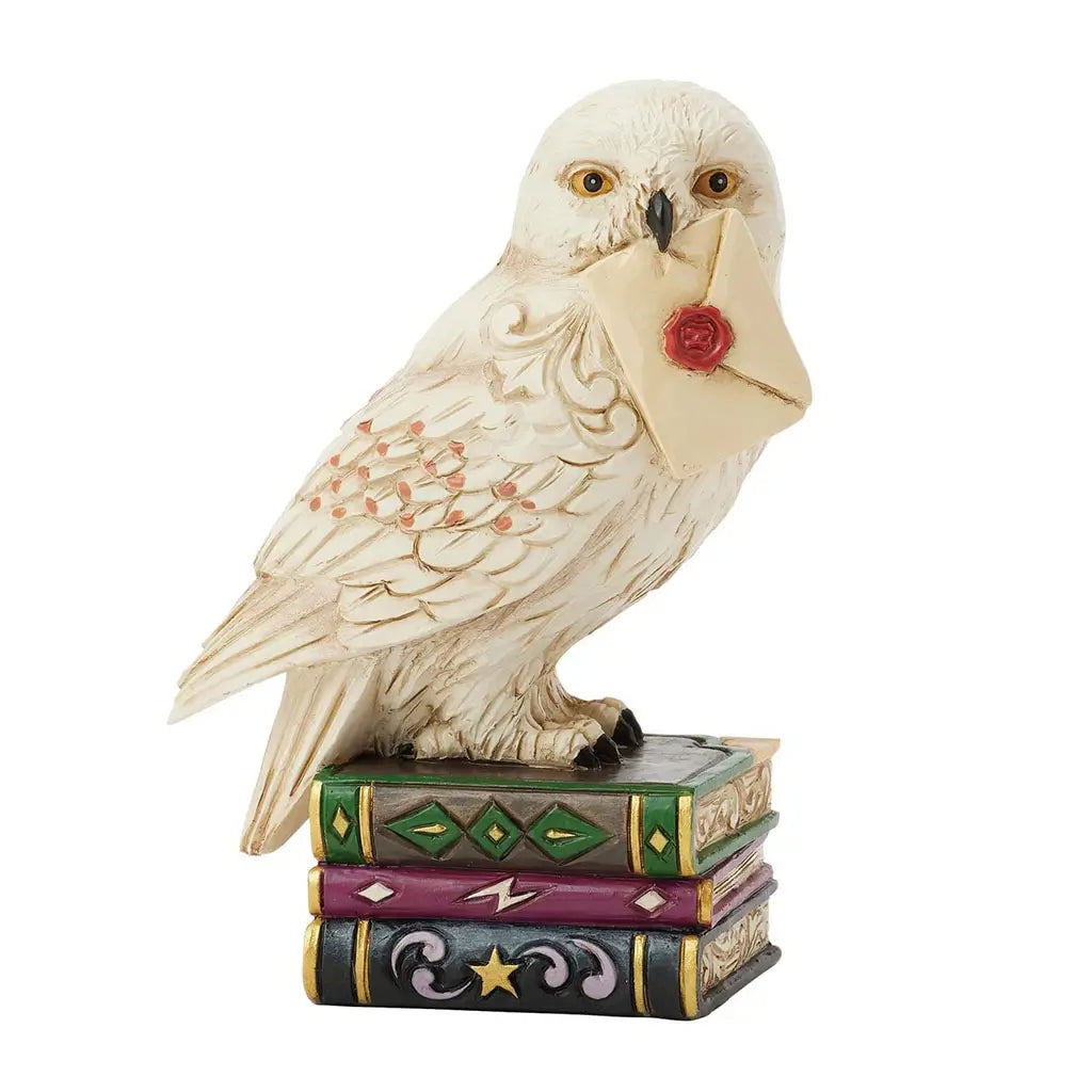 Front view of Jim Shore Hedwig mini figurine depicts owl clutching sealed letter in beak, standing on decorative stack of green, purple, and black books.
