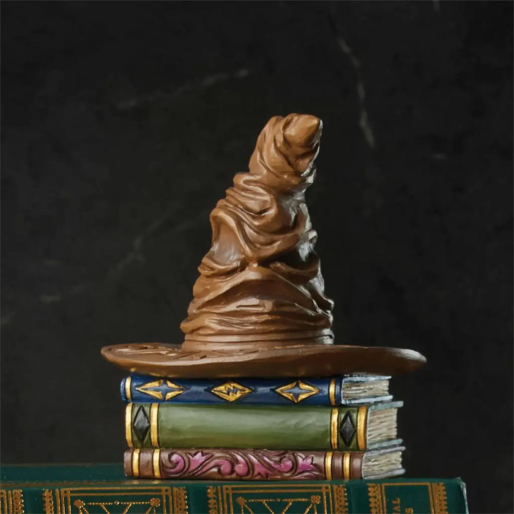 Lifestyle photo of Jim Shore Sorting Hat mini figurine placed on green books, featuring wrinkled brown hat sitting on painted magical book pedestal.