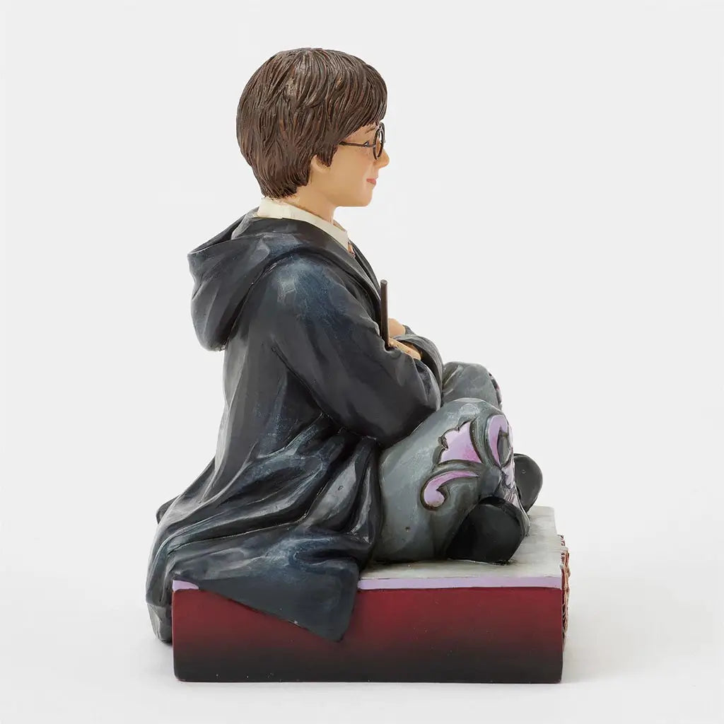 Left side view of Jim Shore Harry Potter figurine, painted in Hogwarts robes, holding a wand, seated on a decorative base with crest carving.