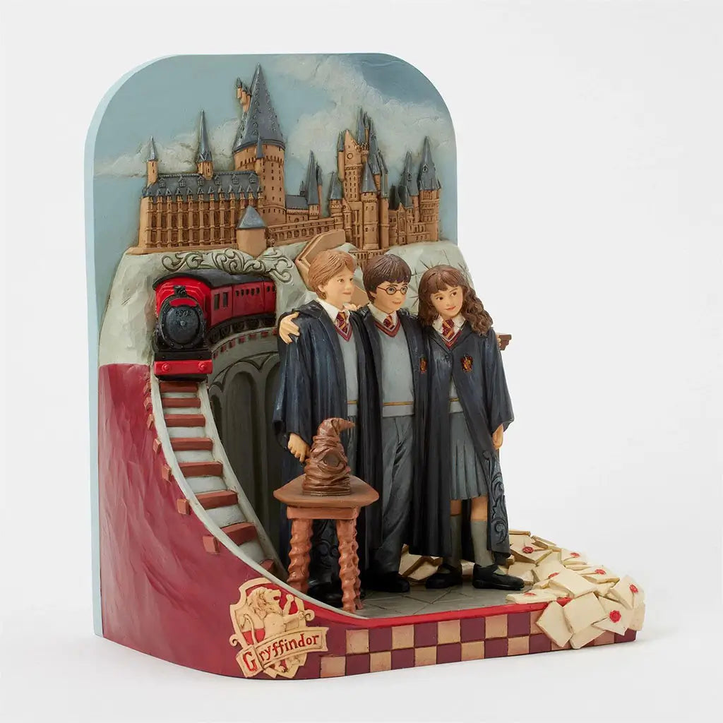 Jim Shore Harry Potter figurine angled left displays trio in Gryffindor robes beside Sorting Hat, Hogwarts Express, castle backdrop, and cascading Hogwarts letters.