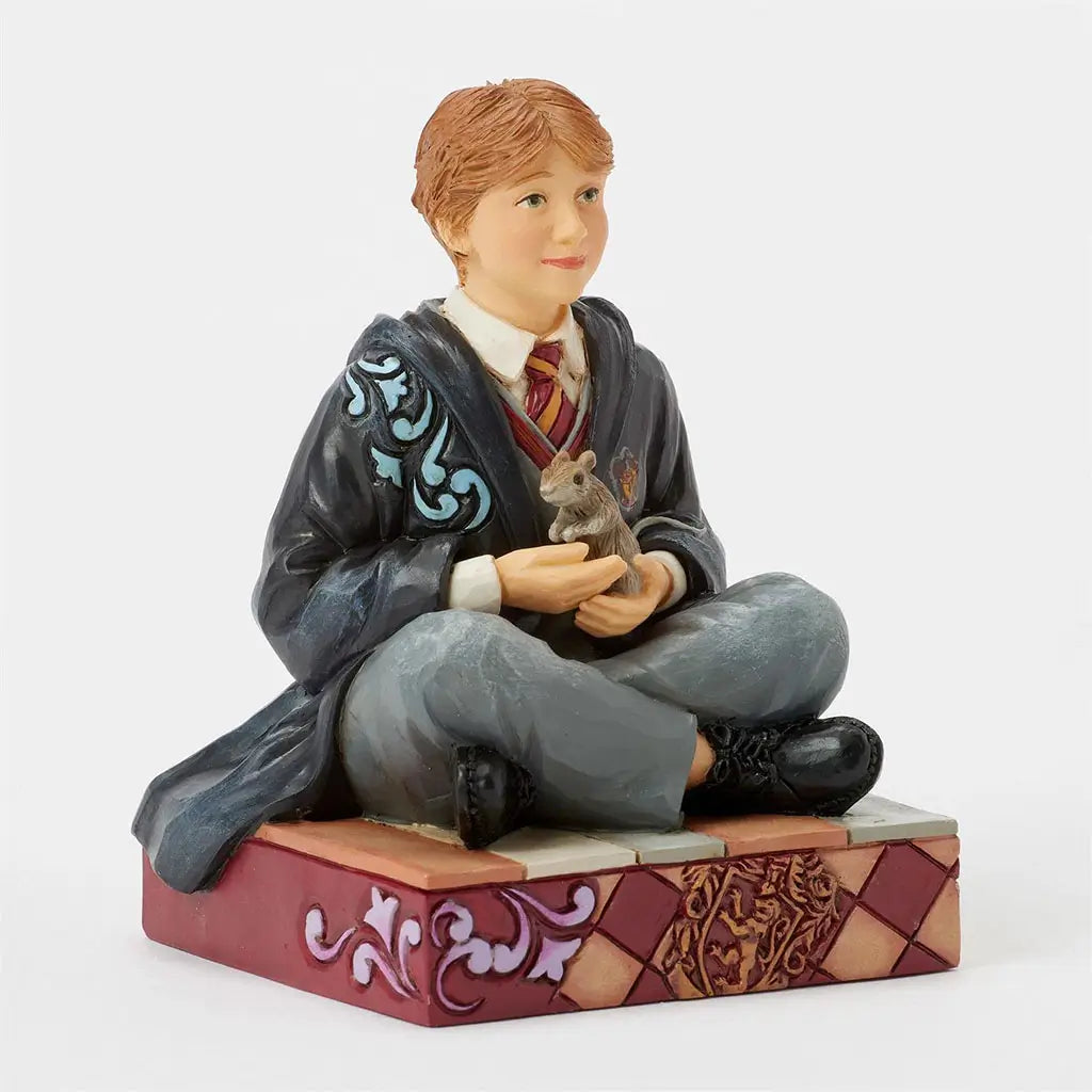 Angled view of Ron Weasley Jim Shore figurine, detailed in red hair, grey pants, black shoes, and maroon tie, holding Scabbers while seated on a decorative platform.