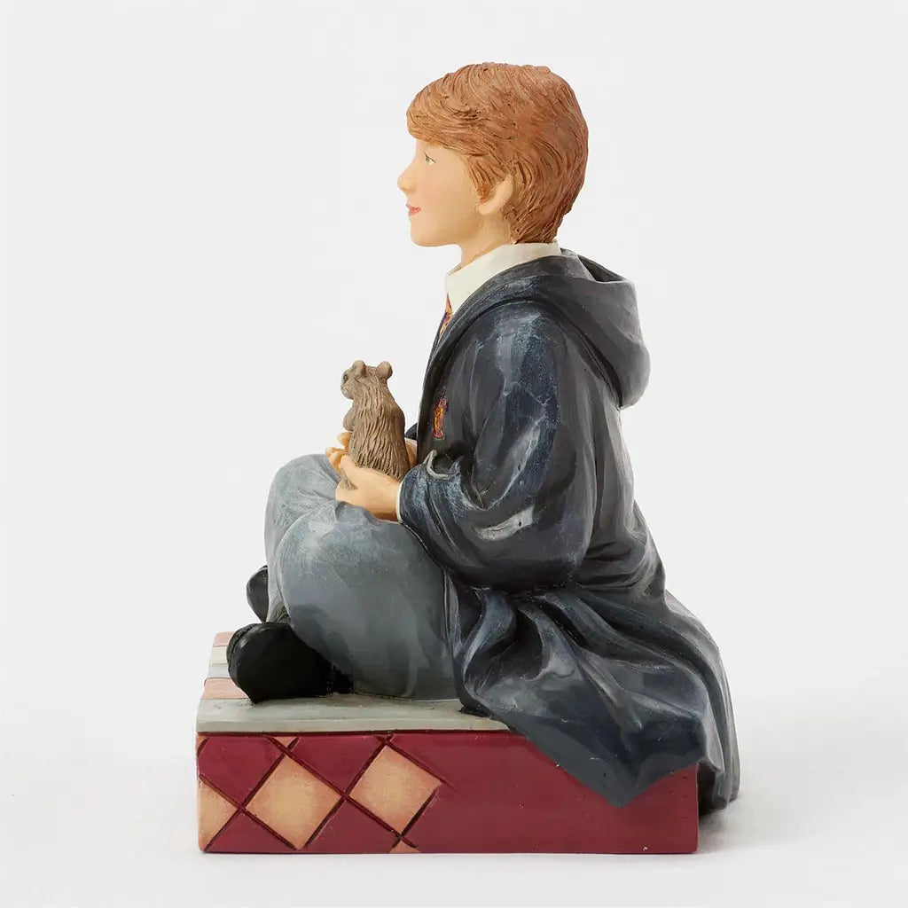 Right side view of Jim Shore Harry Potter figurine of Ron Weasley, hand-painted in Gryffindor robes, sitting cross-legged holding Scabbers the rat on a patterned base.